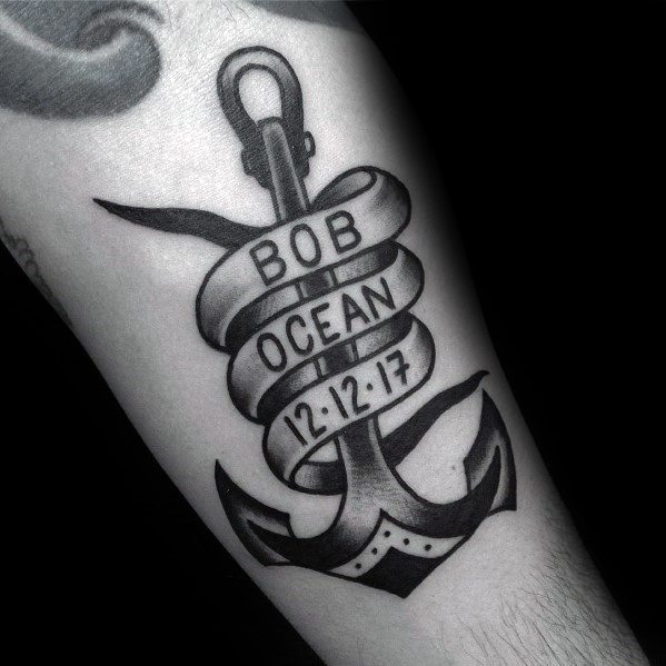 Black and gray anchor tattoo with a ribbon wrapped around it, featuring a name and date, symbolizing remembrance, stability, and personal significance.