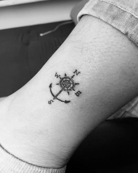 Small anchor and compass tattoo on the ankle, featuring fine black lines and directional markers, symbolizing guidance, stability, and adventure.