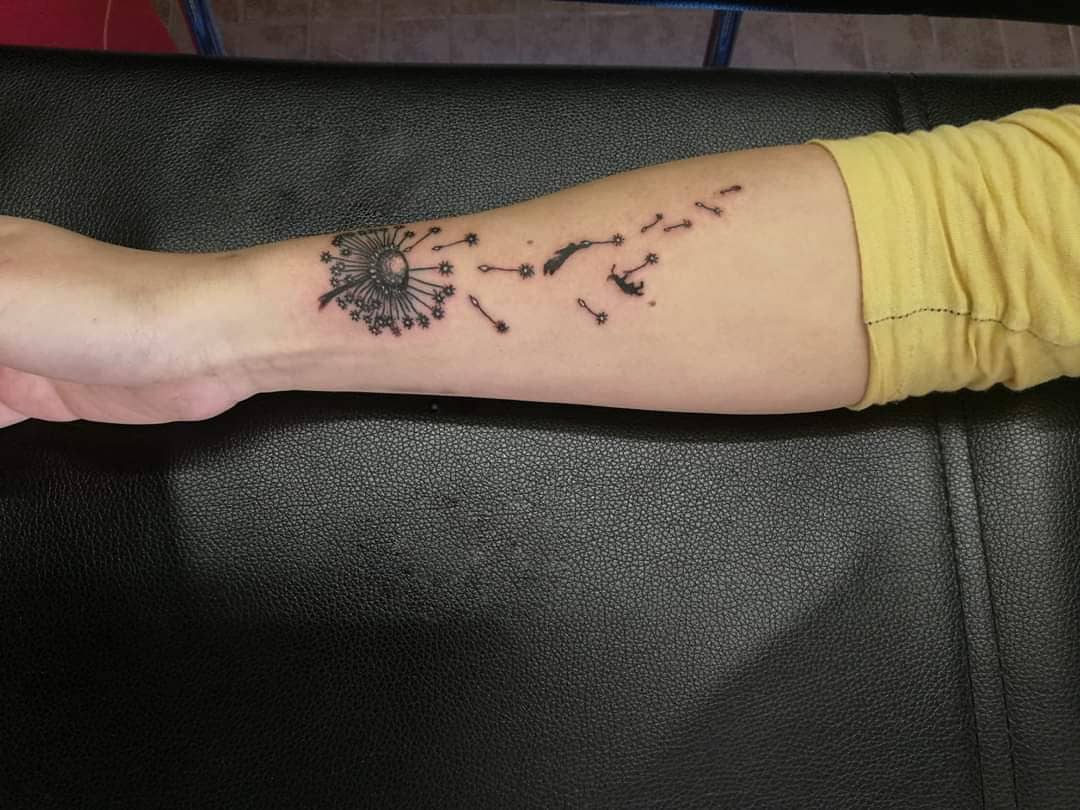 This tattoo depicts a dandelion with seeds dispersing, representing freedom and the fleeting nature of time. It is elegantly designed on the arm with soft lines, allowing the seeds to flow in a graceful, scattered pattern.