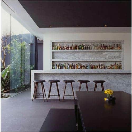 Modern bar featuring a marble counter, five wooden stools, and shelf-lined with bottles; glass wall offers plant views