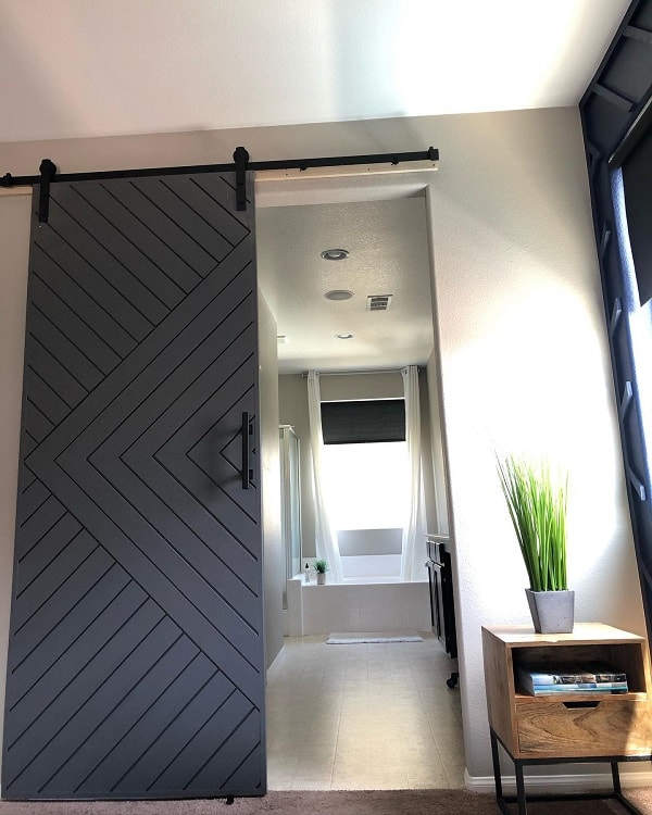 Simple dark gray sliding barn door with chevron pattern leading to a bright bathroom with a bathtub.