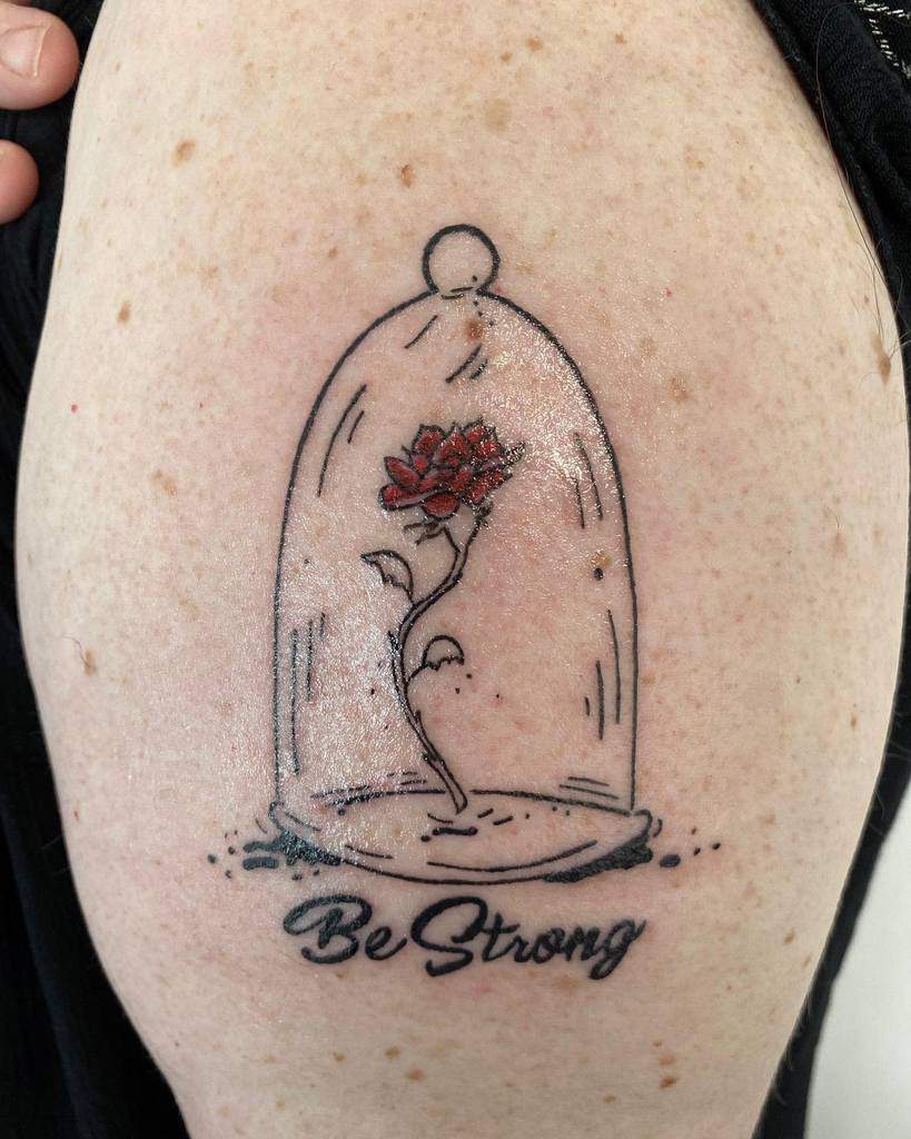 A simple tattoo of the Beauty and the Beast rose inside a glass dome, with a red rose and the motivational phrase "Be Strong" beneath it