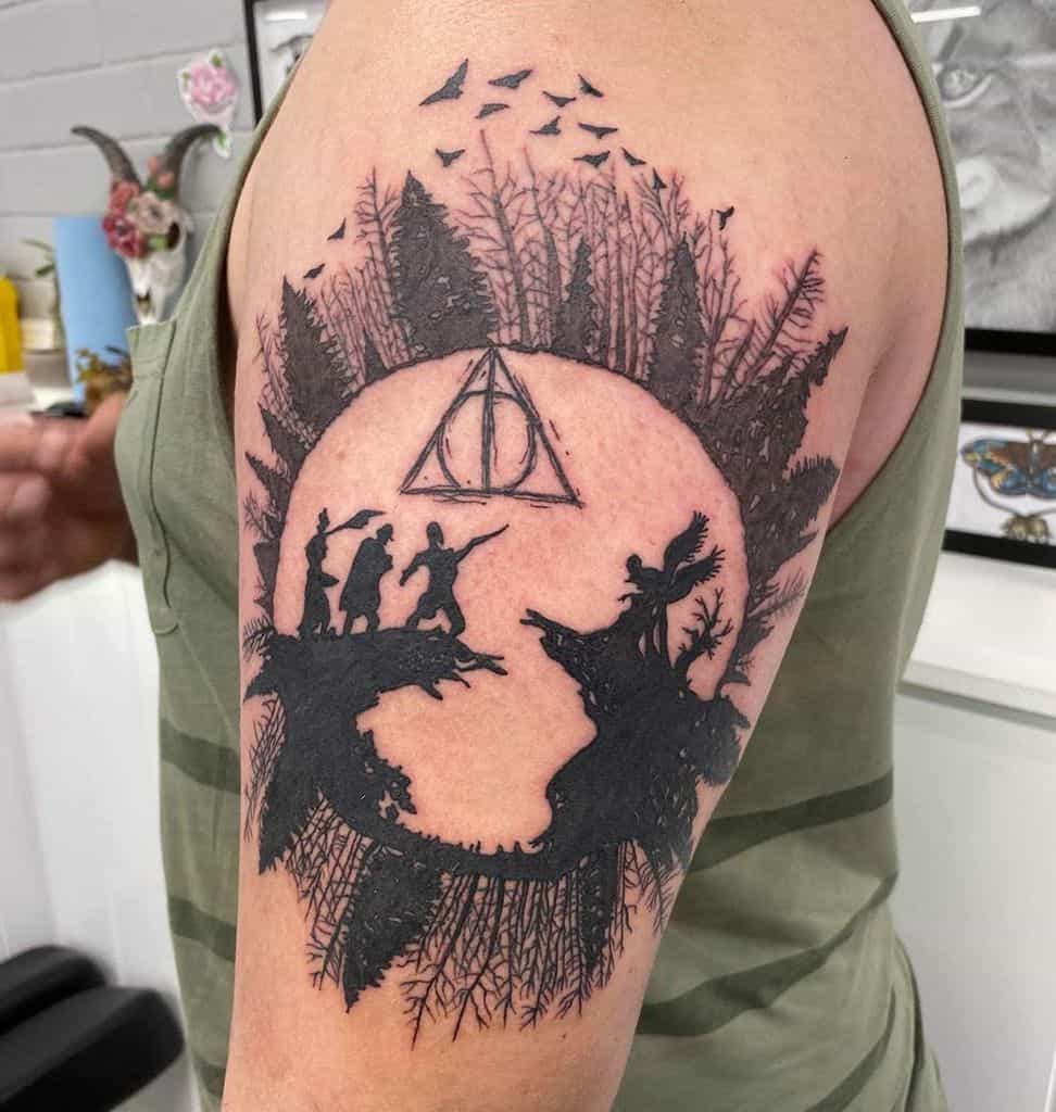Black ink Deathly Hallows tattoo on the upper arm featuring the Tale of the Three Brothers, a forest silhouette, and flying birds.