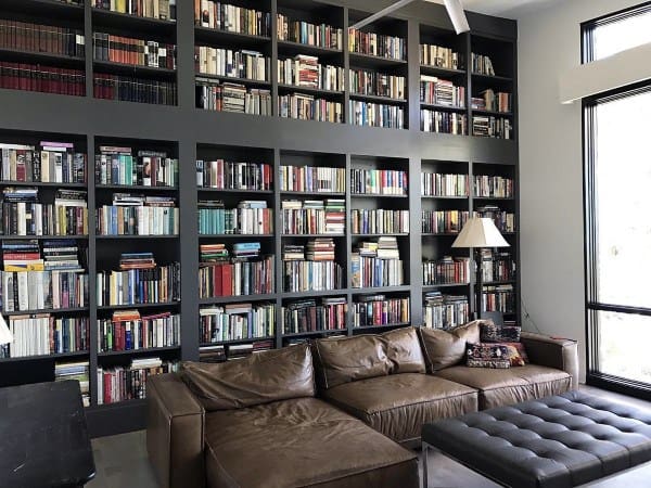 A cozy library with a brown leather sofa, a lamp, and bookshelves filled with books by a large window