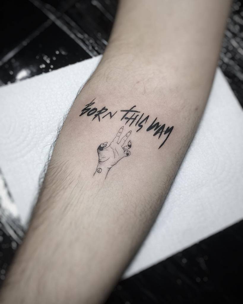 Minimalist tattoo with 'Born This Way' text in bold font above a detailed hand making a peace sign, placed on the forearm