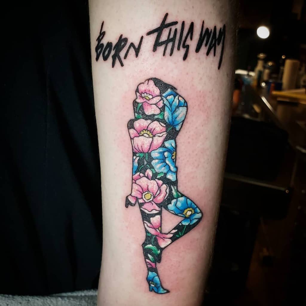 Tattoo of a silhouette filled with colorful flowers under text "Born This Way" on an arm