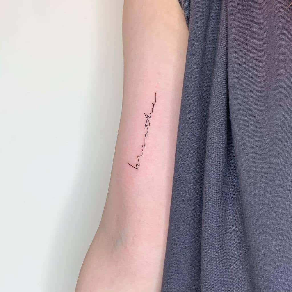 A minimalist tattoo elegantly spells "breathe" in flowing cursive on a person's upper arm, perfectly complementing the subtle gray of their sleeve