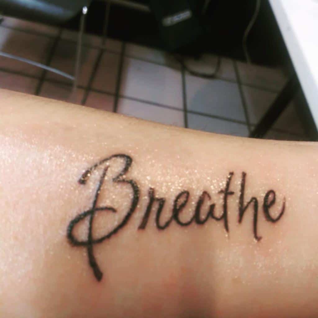 A close-up reveals a delicate tattoo in cursive, the word "Breathe" elegantly etched onto the forearm