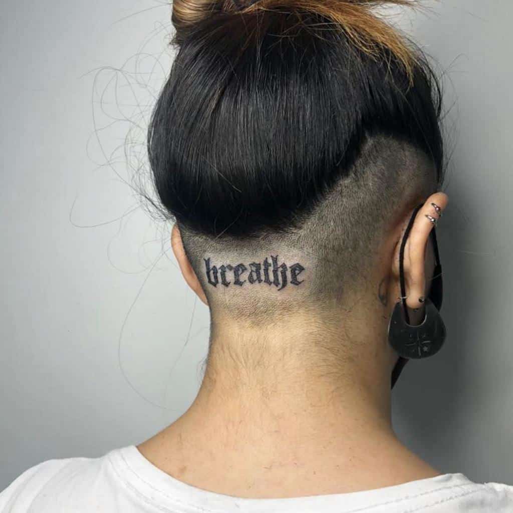 Simple tattoo of the word 'breathe' in gothic font on the back of the neck, placed just below an undercut hairstyle