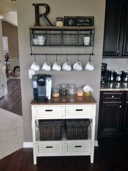 White coffee station with wood countertop, hanging mugs, storage baskets, and metal shelf.