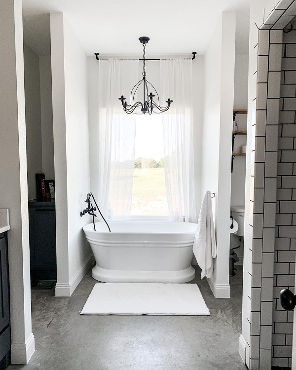 A white freestanding bathtub under a chandelier, with a large window and a white rug on the concrete floor