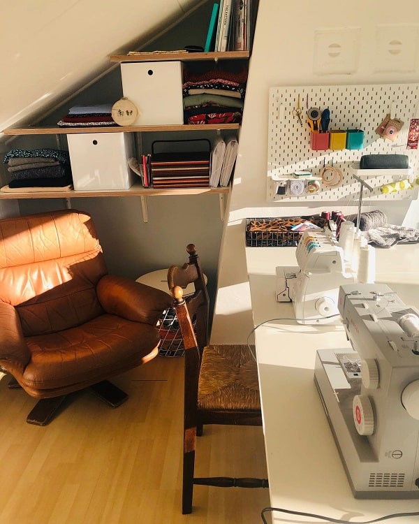 Cozy sewing room with machines, fabric shelves, a pegboard with supplies, and a leather armchair under a sloped ceiling