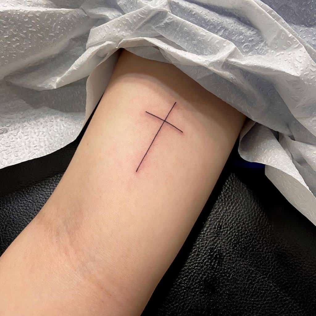 A minimalist black cross tattoo graces the inner forearm