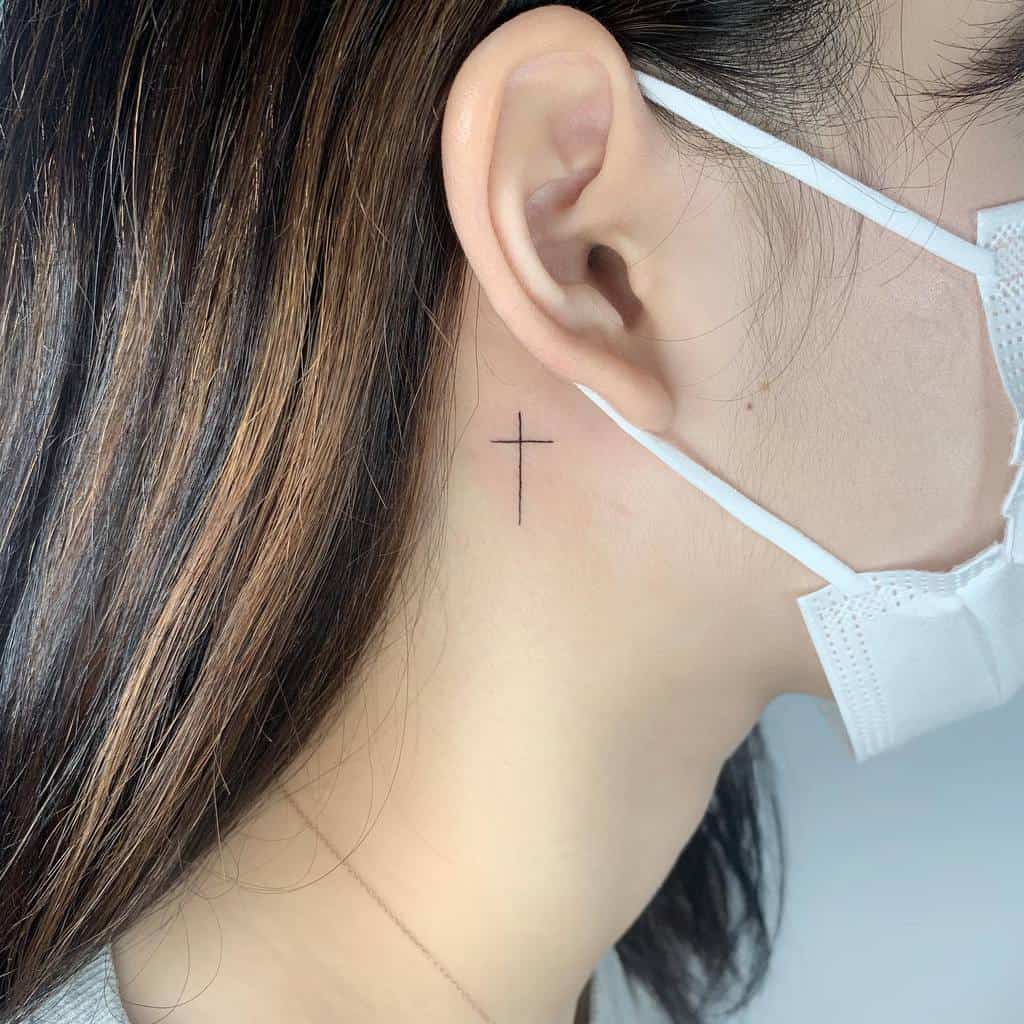 A small and simple cross tattoo located behind the ear, featuring a clean and minimalist design for a subtle yet meaningful look