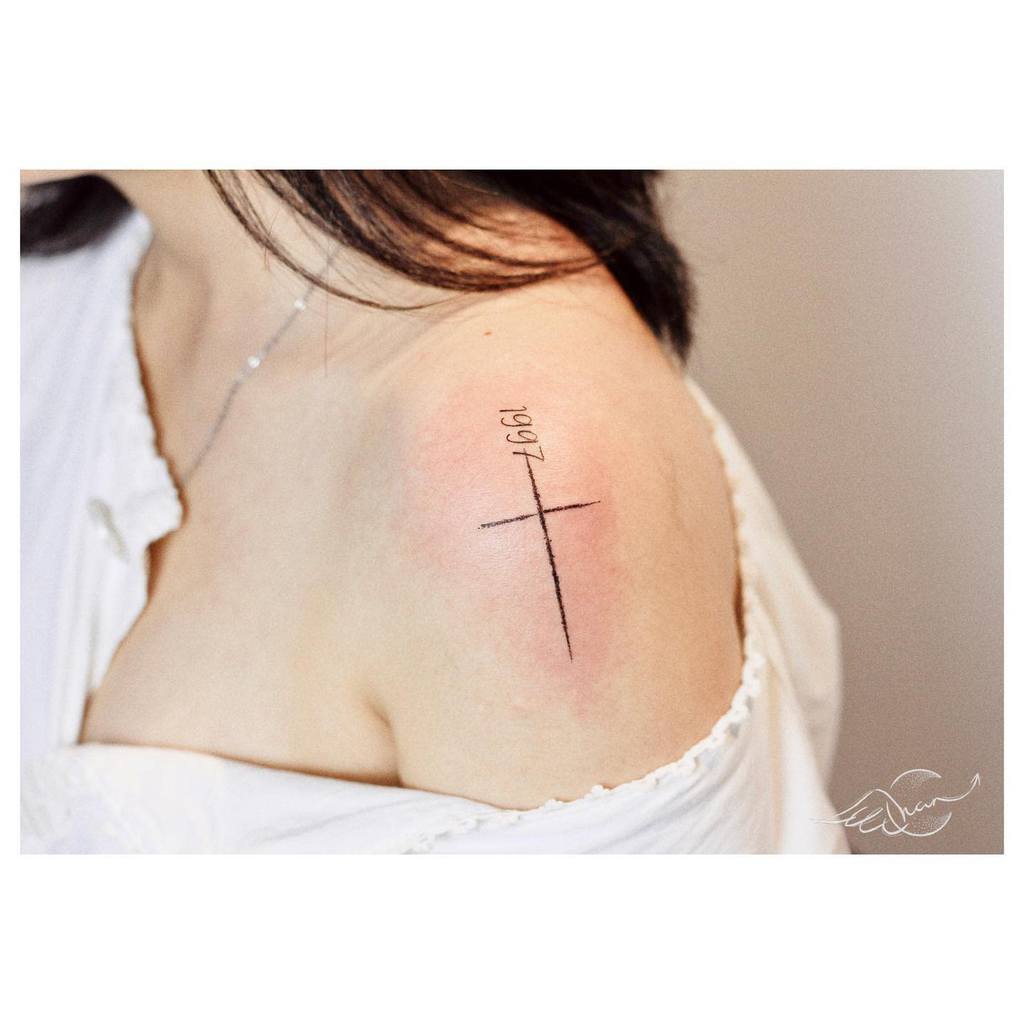 A minimalist cross tattoo featuring "1997" elegantly graces a woman's upper shoulder