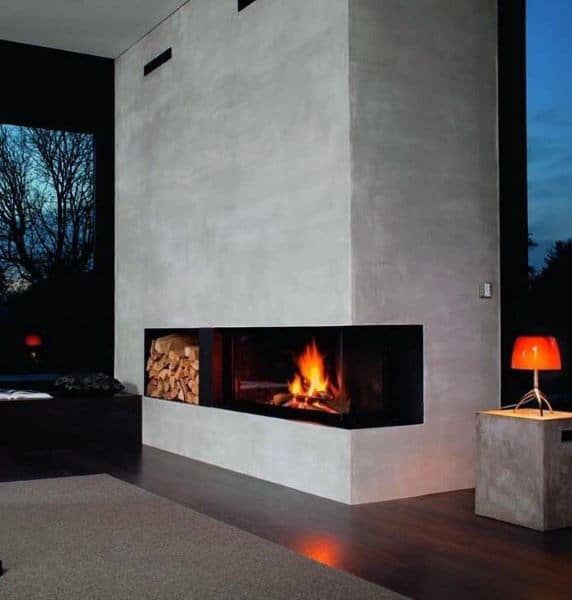Modern corner fireplace with concrete surround, wood storage, and cozy evening lighting.