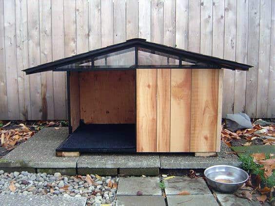 Simple dog house with a glass and metal roof, wooden walls, a small floor mat inside, and a water bowl outside, set on a paved surface