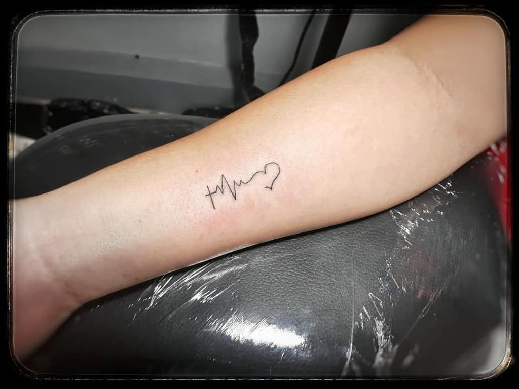 Forearm tattoo with minimalist design featuring a cross, heartbeat line, and heart symbol