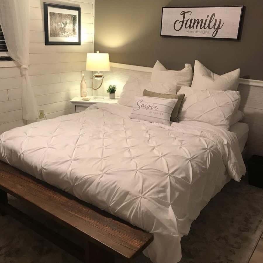 Cozy bedroom with a white comforter, wooden bench, side lamp, and family sign on the wall