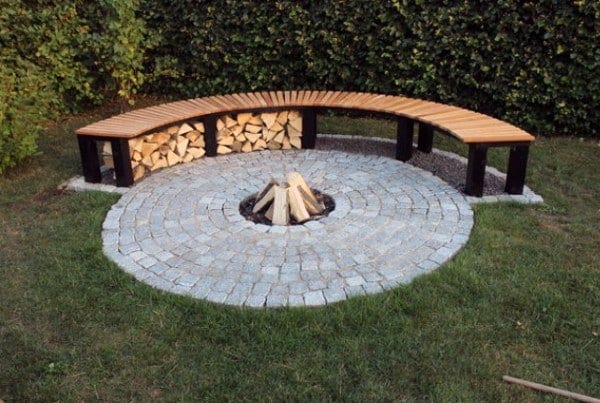 Curved wooden bench by a fire pit on a circular stone patio, surrounded by greenery