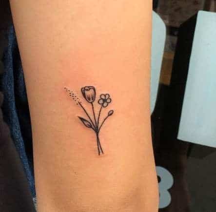 A small black-line tulip tattoo on the arm, featuring a delicate flower with tiny leaves, a small blossom, and subtle decorative dots.
