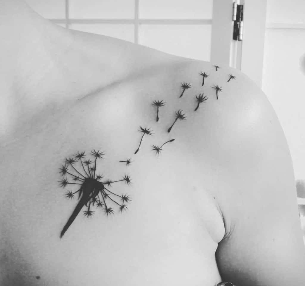 This tattoo features a dandelion on the shoulder with seeds blowing away, symbolizing freedom and transformation in a graceful, minimalist design.