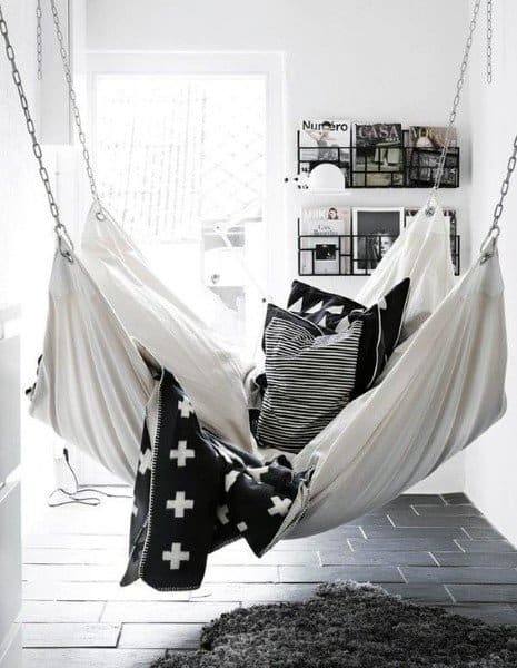 Minimalist hammock-style hanging bed with monochrome cushions and blankets in a bright room.