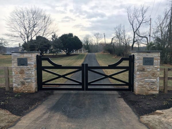 Elegant black driveway gate with a cross-brace design and curved tops, framed by stone pillars with address plaques.
