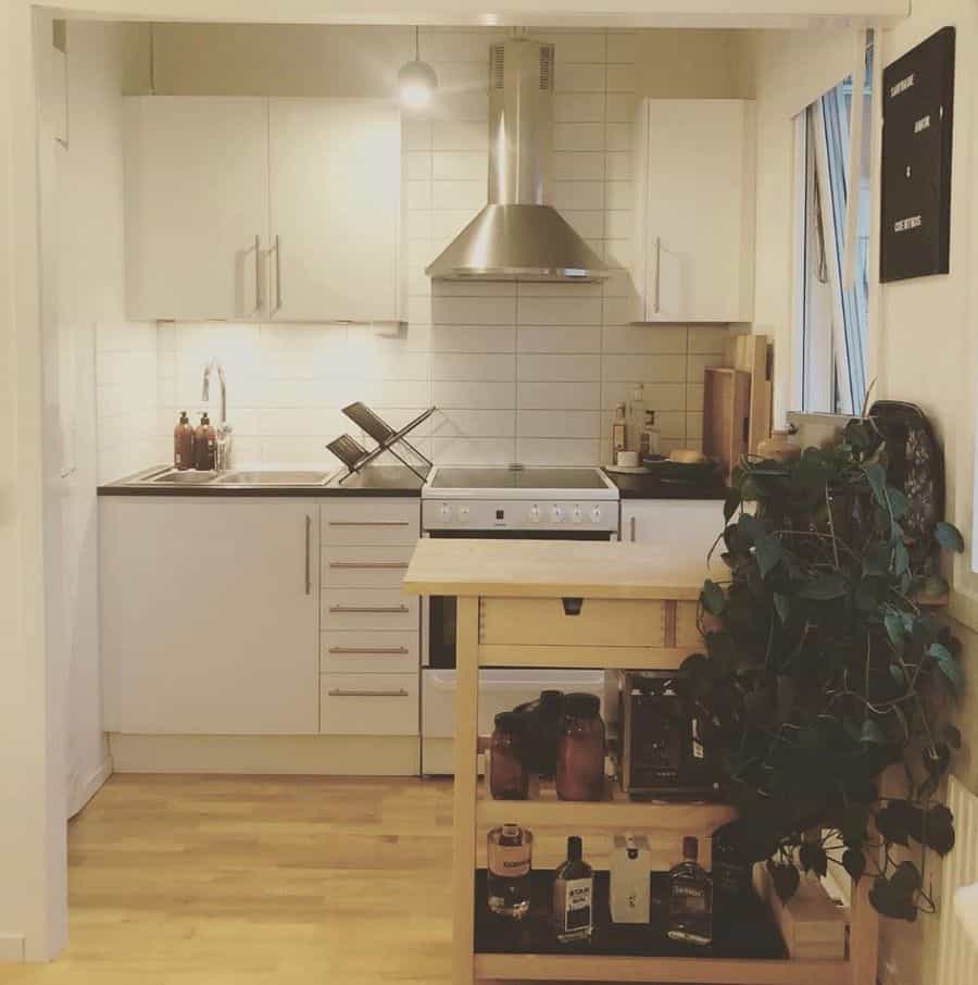Minimalist kitchenette with white cabinetry, subway tile backsplash, and a wooden rolling cart for extra storage.