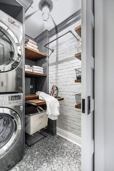 Compact laundry room with stacked appliances, exposed brick, and rustic wooden shelves.