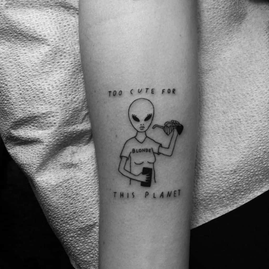 A black ink tattoo of an alien wearing a 'Blonde' shirt, holding a drink and pouring another, with 'Too Cute for This Planet' above and below.