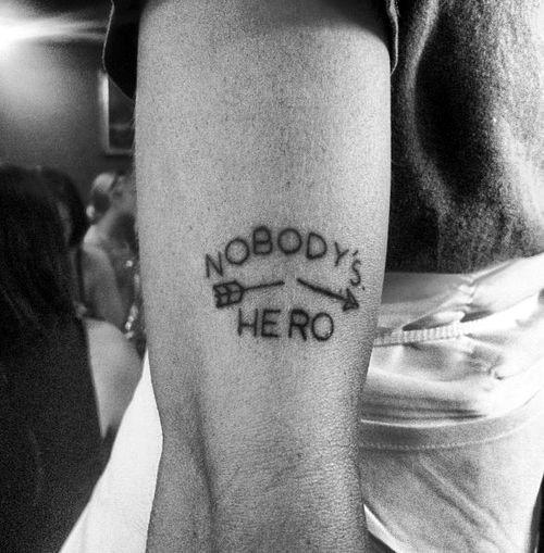 Black ink tattoo featuring the phrase "Nobody's Hero" with a small broken arrow placed between the two words on the back of the arm.