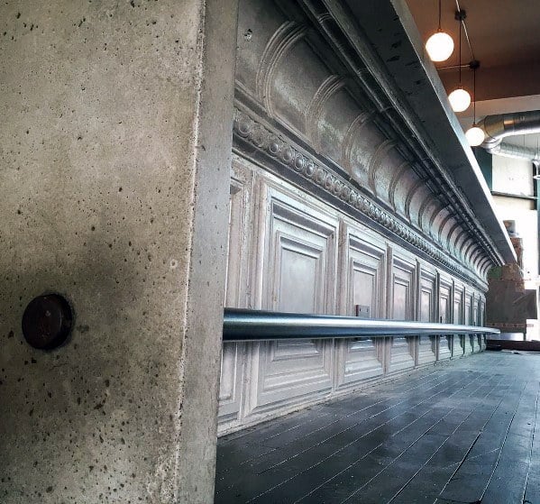 Simple metal foot rail attached to an ornate vintage bar with dark wooden flooring and industrial decor.