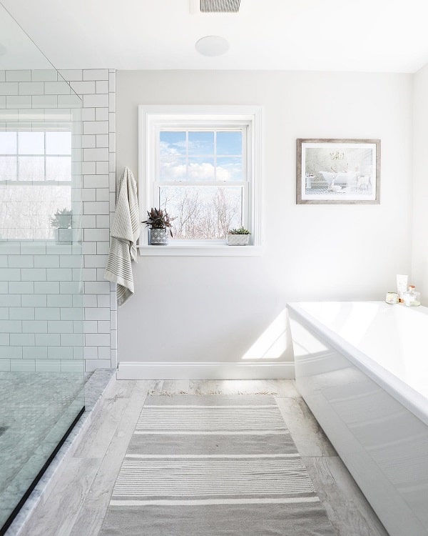 Farmhouse-style bathroom with a glass shower, window, bath, and gray-striped rug, featuring towels hanging near the subway-tiled shower