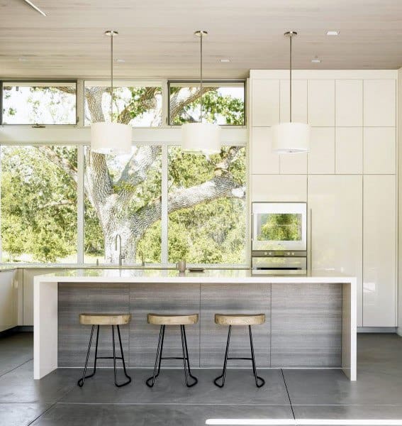 Minimalist kitchen with a white island, wooden stools, and large windows framing greenery.