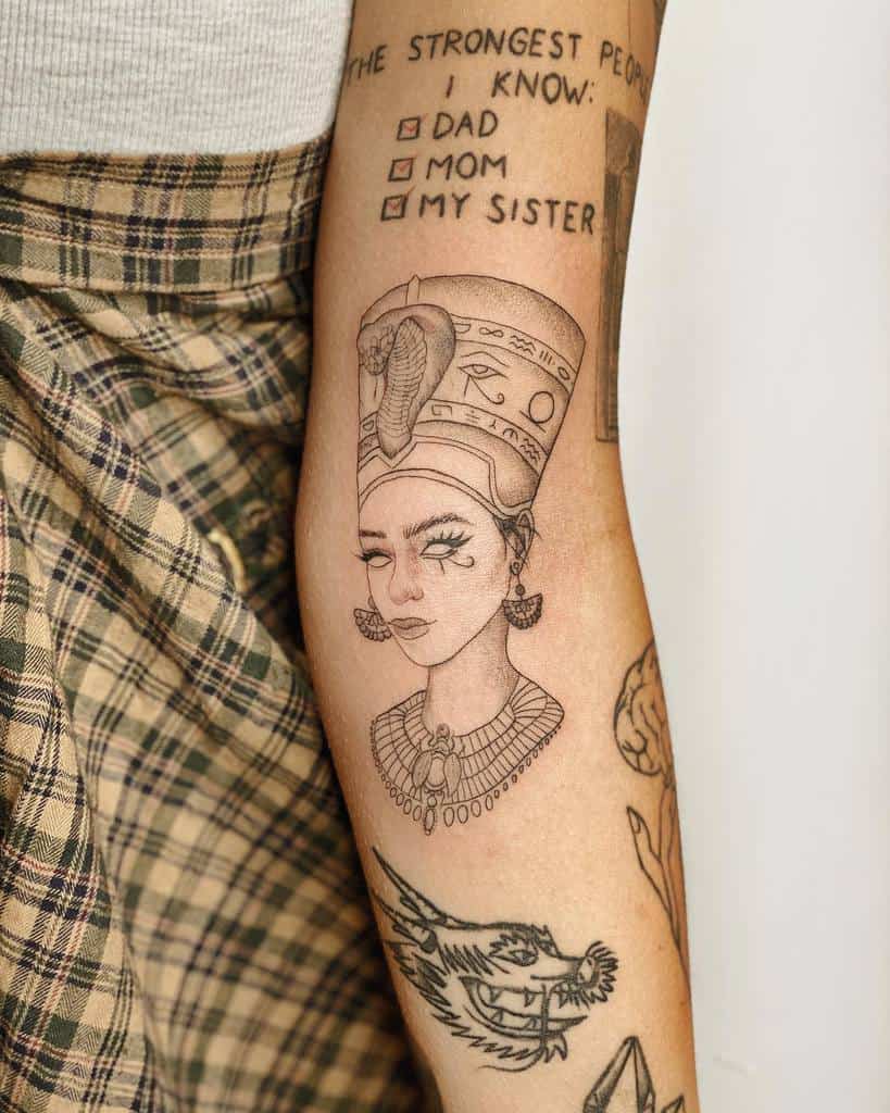 Detailed forearm tattoo of Queen Nefertiti in fine linework, surrounded by other tattoos with meaningful text and artistic designs