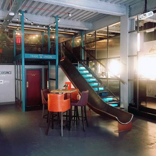 Modern office featuring a slide and turquoise-lit stairs, surrounded by a high table with orange chairs