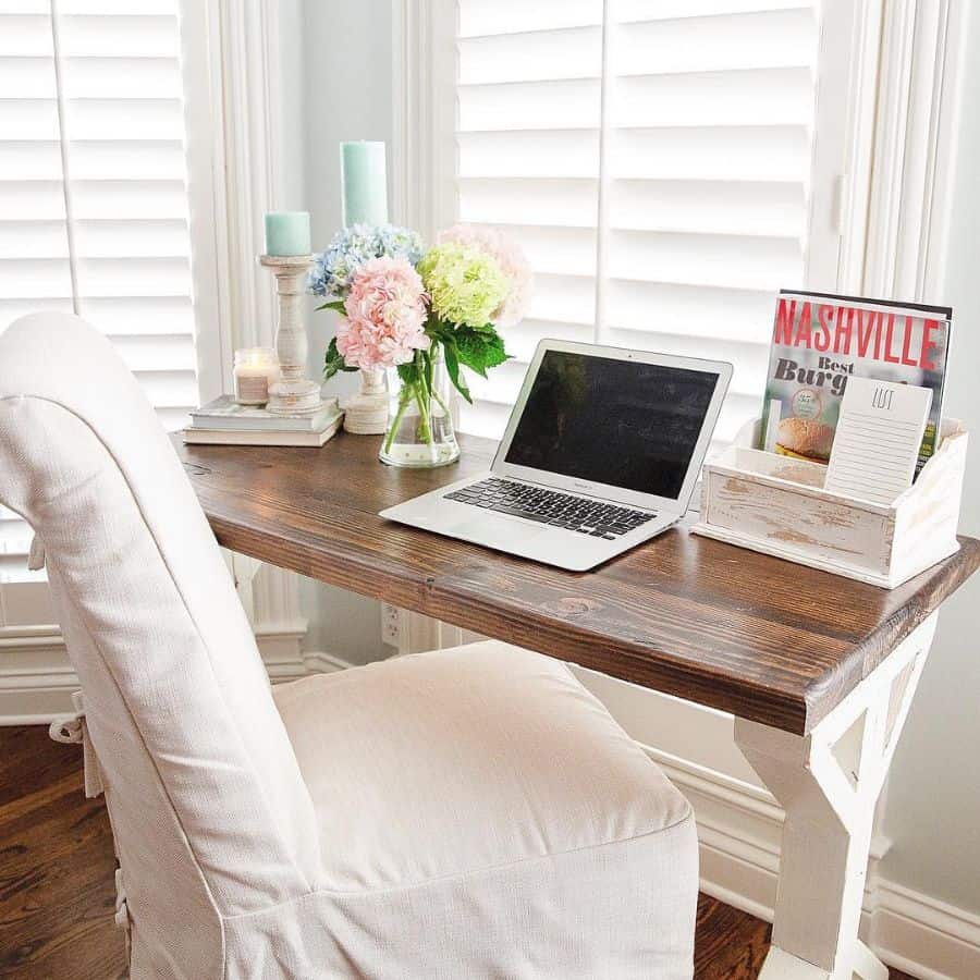 Cozy workspace with wooden desk, laptop, flowers, and decorative accents by a window.
