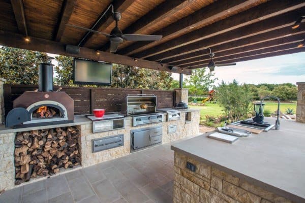 Outdoor kitchen with wood-fired oven, grill, and prep area under a wooden pergola, overlooking an outdoor garden