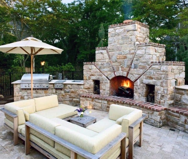 Cozy patio with beige seating, a brick fireplace, grill, and umbrella, nestled in lush greenery