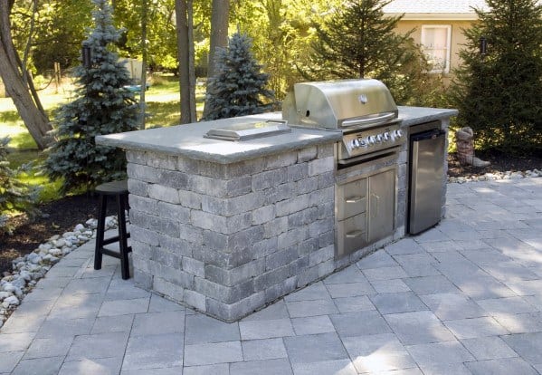 Outdoor kitchen with stone counter, built-in grill, and mini fridge on a paved patio enveloped by trees