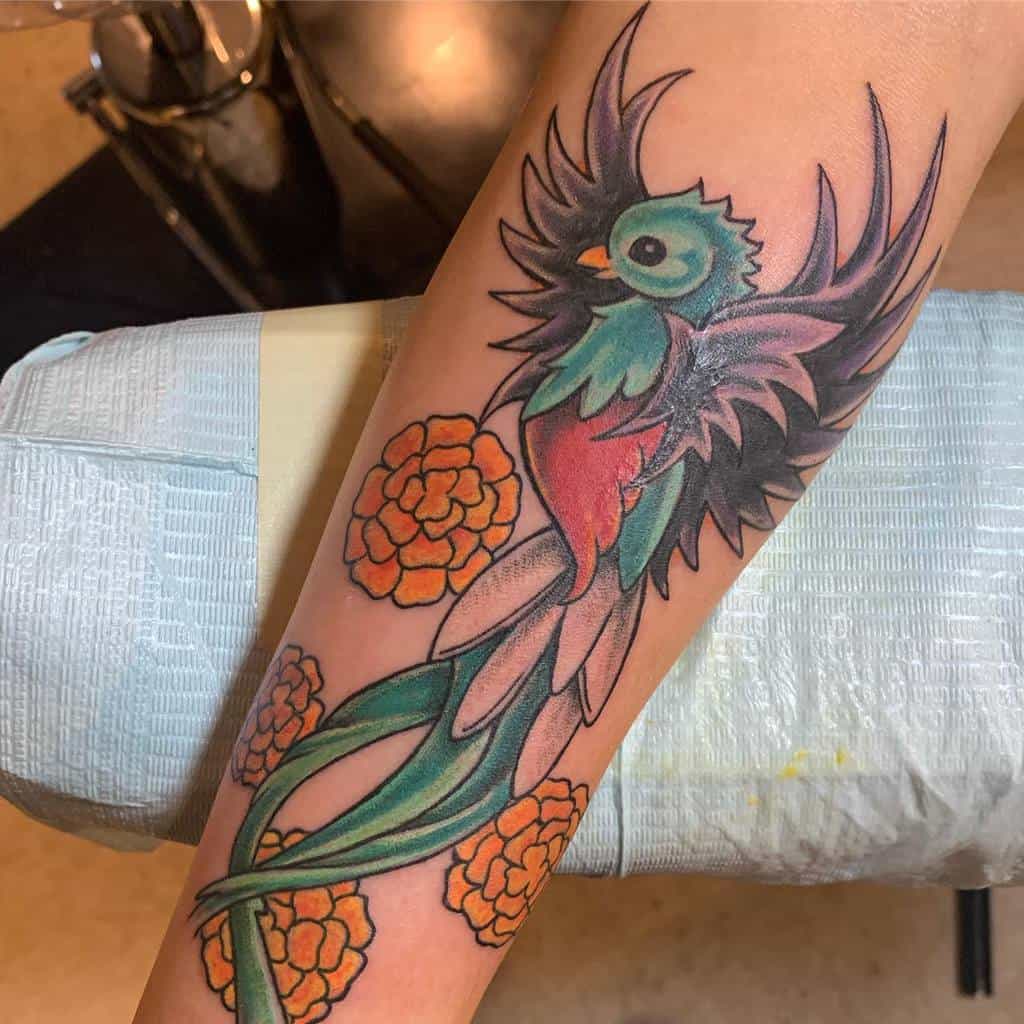 Bold quetzal tattoo on the forearm, featuring a stylized green and red bird with dramatic black wings, flowing tail feathers, and vibrant orange flowers