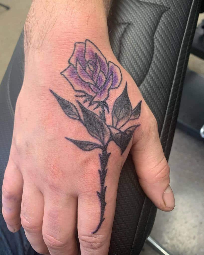 A tattoo of a vibrant purple rose blooms gracefully with lush leaves on the back of a hand