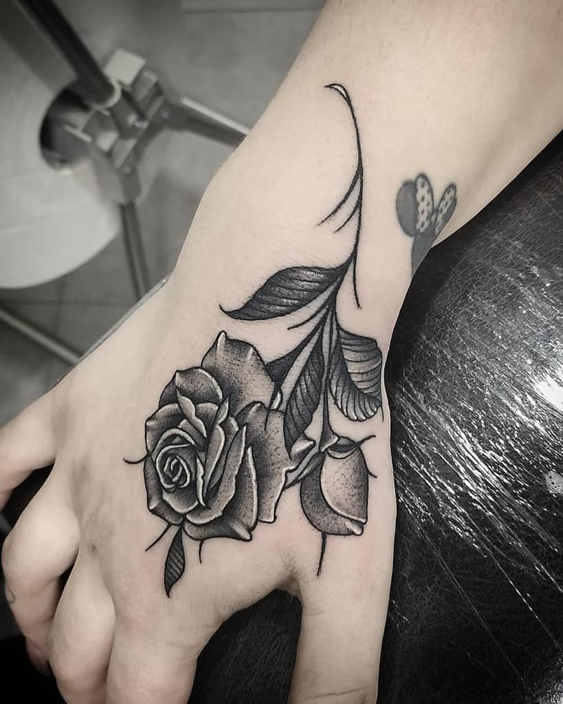 A stunning black and gray tattoo adorns a person's hand, featuring a beautifully detailed rose with leaves and intricate small accents
