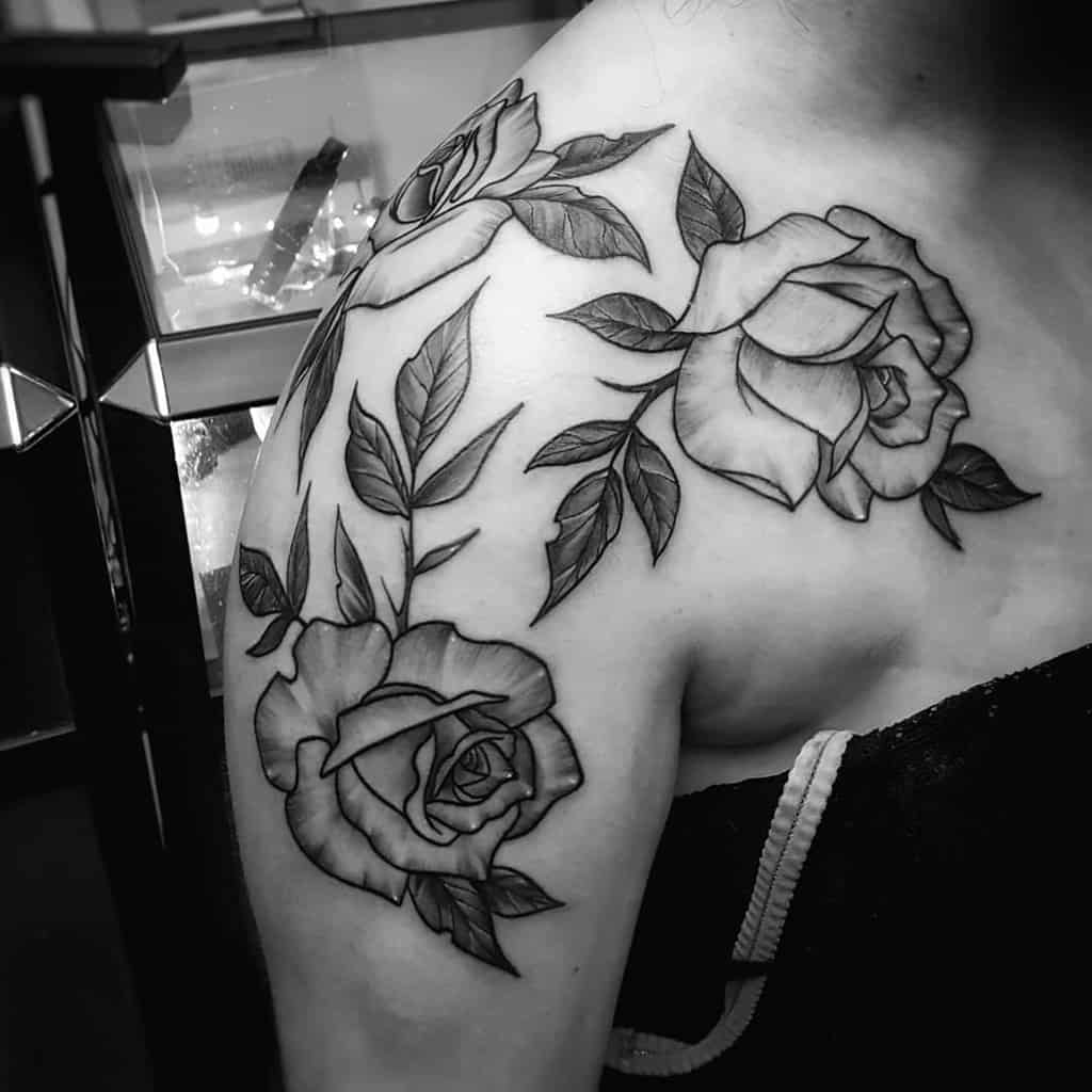 A black and grey rose tattoo on the shoulder, featuring three roses surrounded by leaves, with soft shading and clean outlines
