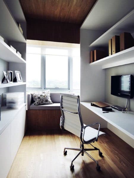 Minimalist home office with white shelves, desk, and chair; a window seat with cushions is by the large window