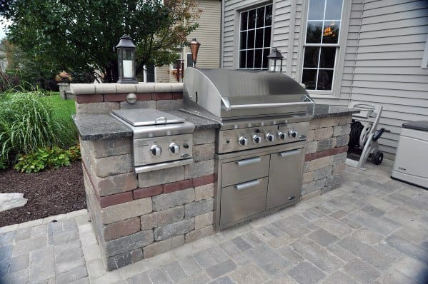 Outdoor barbecue station with built-in grill and stainless steel appliances, set on a stone patio beside a house