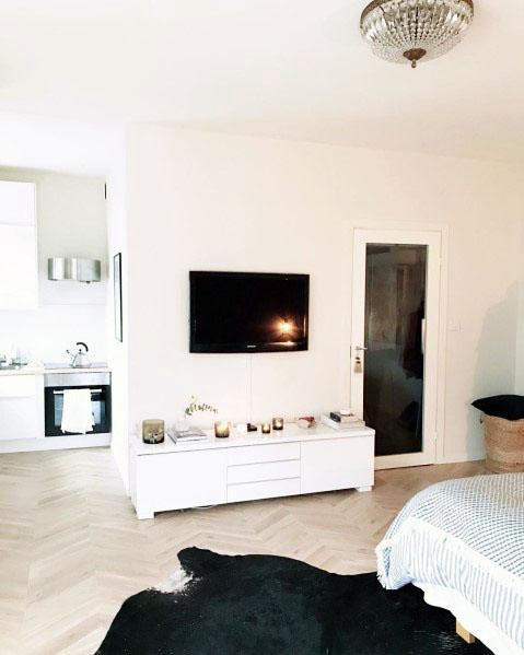 Minimalist studio with white decor, wall-mounted TV, compact kitchen, and herringbone flooring.