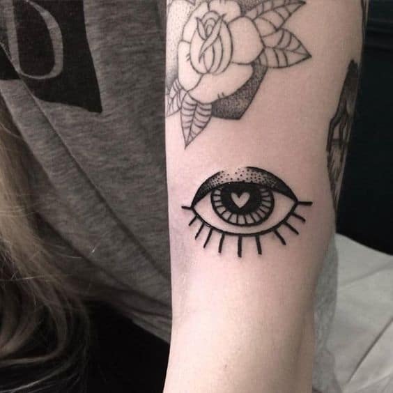 Tattoo of a stylized all-seeing eye with bold black shading and a small heart in the pupil, inked on the arm.