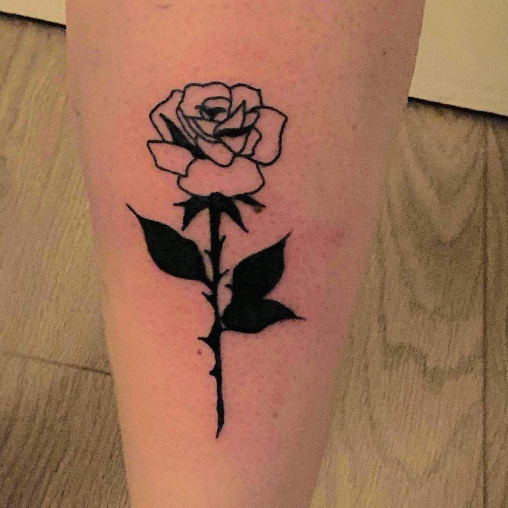 Tattoo of a black rose with a long stem and leaves on a leg